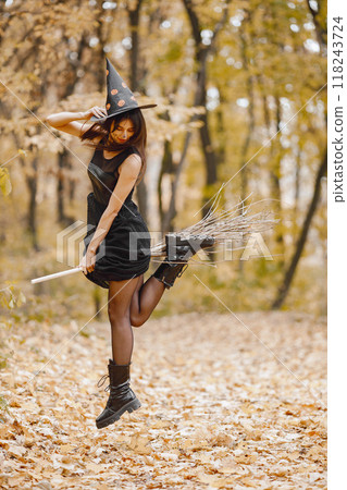 Brunette young girl witch in forest on Halloween. Girl wearing black dress and cone hat. Witch holding a magician stuff. 118243724