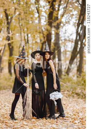 Three young girls witches in forest on Halloween. Girls wearing in black dresses and cone hat. Witch holding a magician stuff. Three young girls witches in forest on Halloween. Girls wearing in black dresses and cone hat. Witch holding a magician stuff. 118243727