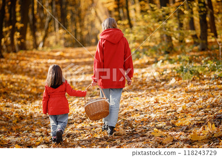 Young woman with little girl walking in autumn forest. Blonde woman play with her daughter and holding a basket. Mother and daughter wearing jeans and red jackets. Young woman with little girl walking in autumn forest. Blonde woman play with her daughter and holding a basket. Mother and daughter wearing jeans and red jackets. 118243729