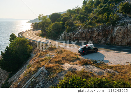 Black car with a paddleboard strapped to the roof, parked at a cliffside viewpoint with azure sea below Black car with a paddleboard strapped to the roof, parked at a cliffside viewpoint with azure sea below 118243925