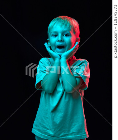 Portrait of cute little boy, child in casual white t-shirt holding hand to cheeks and smiling against black studio background in neon light Portrait of cute little boy, child in casual white t-shirt holding hand to cheeks and smiling against black studio background in neon light 118245373
