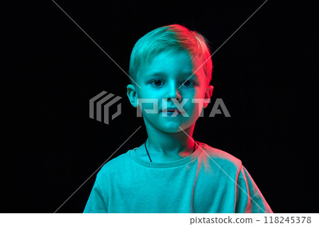 Portrait of adorable little baby boy, blond haired child wearing t-shirt and looking at camera with warm smile against black studio background in neon light 118245378