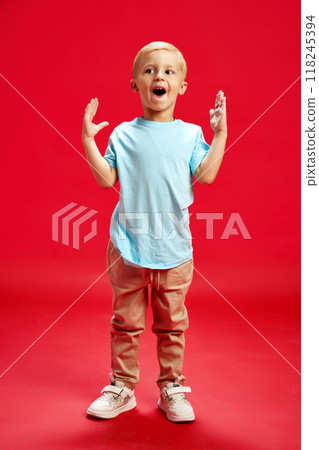 Full-length portrait of joyful little boy, child in casual clothes, t-shirt and pant design with joy and fun against red studio background 118245394