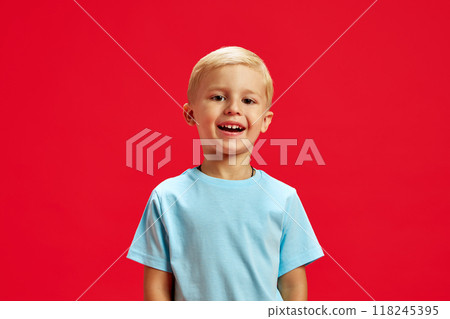 Portrait of little 4 years old boy with blonde hair, standing with l smile, wearing blue t-shirt and laughing against red studio background 118245395