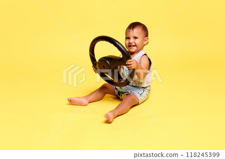 Beautiful little baby, girl sitting in a denim outfit, holding steering wheel, smiling with pure joy against yellow studio background 118245399