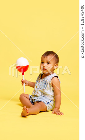 Beautiful little baby, girl sitting in a denim outfit, holding bright toy, completely immersed in world of exploration against yellow studio background Beautiful little baby, girl sitting in a denim outfit, holding bright toy, completely immersed in world of exploration against yellow studio background 118245400