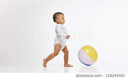 Little baby, girl in onesie walking confidently towards colorful ball, playing with curiosity and joy isolated on white studio background Little baby, girl in onesie walking confidently towards colorful ball, playing with curiosity and joy isolated on white studio background 118245401