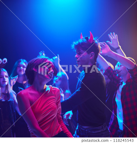 Focus on cheerful young boy and girl dresses like devils and creepy characters, attending Halloween themed party and dancing, with blurred friends on background 118245543