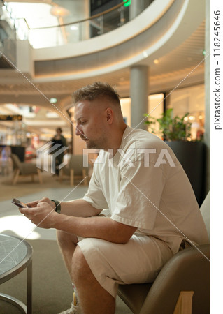 Profile of a happy guy in stylish casual clothes using a smart phone sitting on a soft chair at shopping mall Profile of a happy guy in stylish casual clothes using a smart phone sitting on a soft chair at shopping mall 118245646