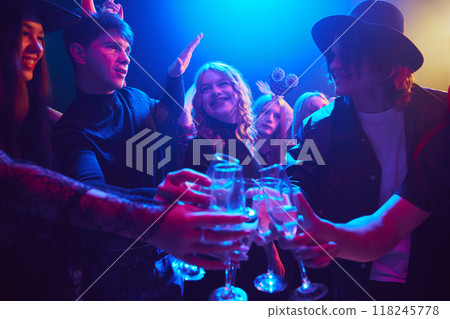 Friends, boys and girls in various Halloween costumes gathering together at nightclub for celebration, clinking glasses with drinks and showing cheerful mood Friends, boys and girls in various Halloween costumes gathering together at nightclub for celebration, clinking glasses with drinks and showing cheerful mood 118245778
