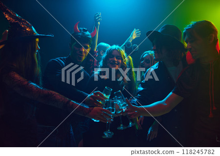 Friends, boys and girls in various Halloween costumes gathering together at nightclub for celebration, clinking glasses with drinks and showing cheerful mood Friends, boys and girls in various Halloween costumes gathering together at nightclub for celebration, clinking glasses with drinks and showing cheerful mood 118245782