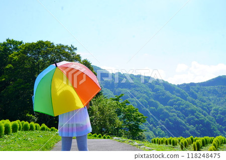 一個女孩在陽光明媚的夏日高原上撐著彩色遮陽傘的後視圖。 一個女孩在陽光明媚的夏日高原上撐著彩色遮陽傘的後視圖。 118248475