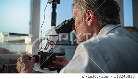 Mature microbiologist analyzes fossil sample under microscope, looks at laptop screen Mature microbiologist analyzes fossil sample under microscope, looks at laptop screen 118248838