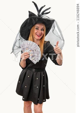 Happy witch in a Halloween costume smiles, gives a thumbs up, and holds a fan of money, exuding positivity and excitement against a white background Happy witch in a Halloween costume smiles, gives a thumbs up, and holds a fan of money, exuding positivity and excitement against a white background 118248894