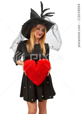 Smiling woman wearing a witch hat and holding a red heart shaped pillow is posing on a white background 118249084