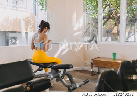 Lazy woman in Yellow Blue fitness outfit sitting and using mobile in the gym. 118249232