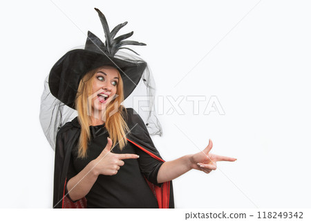 Laughing young woman in black witch hat pointing her fingers to the side. Halloween party, white background, copy space 118249342