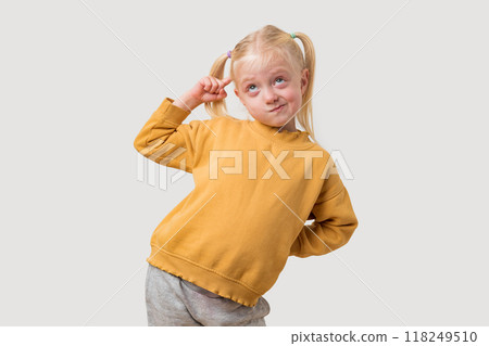 Portrait of a little blonde girl in a yellow sweatshirt puts her finger to her head and thinks on a white background. Portrait of a little blonde girl in a yellow sweatshirt puts her finger to her head and thinks on a white background. 118249510