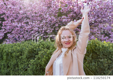 Young beautiful woman smiling and standing in blooming sakura park raised her hands. Happy gentle woman in blossoming park. Young beautiful woman smiling and standing in blooming sakura park raised her hands. Happy gentle woman in blossoming park. 118250174