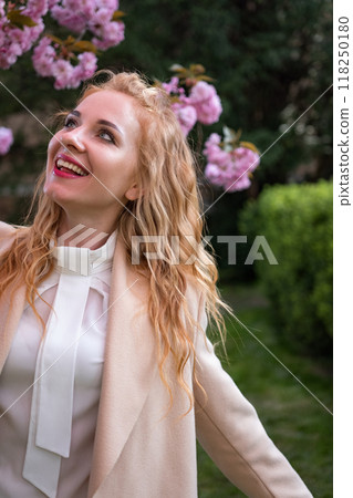 Happy young redhead woman wearing stylish outfit and smiles near blossoming sakura in park. Fashionable spring look. Vertical frame. 118250180