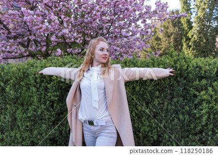 Red haired woman with curly hair raised arms in blooming spring park. Attractive girl on blooming sakura tree background. 118250186