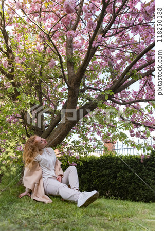 Girl resting in spring park under flowering tree. Young woman sits on the grass under cherry blossoms and looks up. Vertical frame. Girl resting in spring park under flowering tree. Young woman sits on the grass under cherry blossoms and looks up. Vertical frame. 118250188