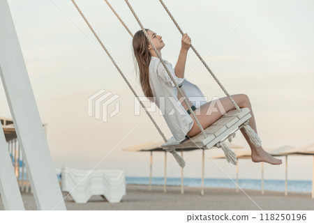 Happy beautiful young girl in white clothes pushes in swing on sky background. Beautiful soft sunset light. Pretty girl on the beach Happy beautiful young girl in white clothes pushes in swing on sky background. Beautiful soft sunset light. Pretty girl on the beach 118250196