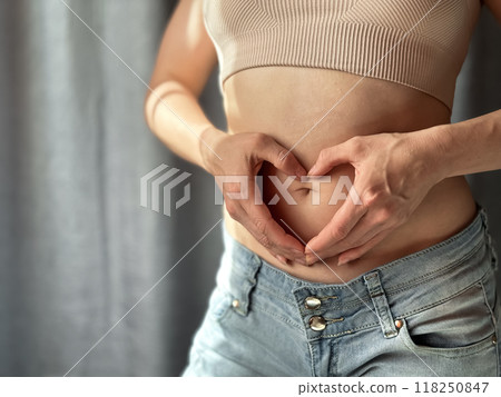 Close up of slim woman in short top and jeans forming heart shape with her hands on her bare stomach. Concept of body positivity, self love, and health awareness. Lifestyle photo. 118250847