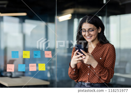 Young business woman using phone inside office, company worker smiling typing text message and browsing social networks, hispanic woman satisfied with work standing near window. Young business woman using phone inside office, company worker smiling typing text message and browsing social networks, hispanic woman satisfied with work standing near window. 118251977