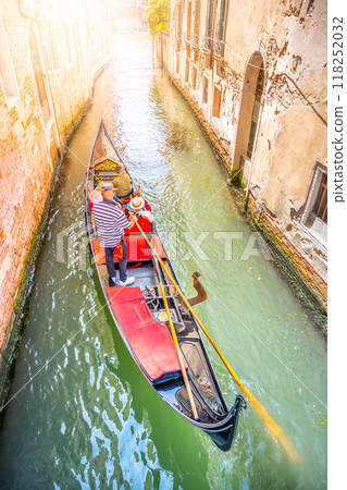 A gondola glides through a peaceful canal in Venice, Italy, as the sun casts a warm glow. The gondolier navigates while passengers enjoy the picturesque surroundings of historic buildings. 118252032