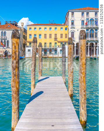 A charming wooden jetty extends into the tranquil waters of Venice, Italy, surrounded by historic buildings and a clear blue sky, inviting leisurely strolls and peaceful moments by the water. A charming wooden jetty extends into the tranquil waters of Venice, Italy, surrounded by historic buildings and a clear blue sky, inviting leisurely strolls and peaceful moments by the water. 118252036