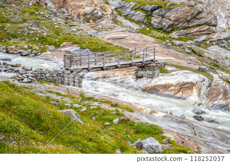 A wooden bridge spans a rushing stream sourced from the Schlaten Glacier, surrounded by rocky terrain and vibrant green vegetation in Hohe Tauern National Park. A wooden bridge spans a rushing stream sourced from the Schlaten Glacier, surrounded by rocky terrain and vibrant green vegetation in Hohe Tauern National Park. 118252037