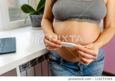An expectant mother in a serene home setting. She gently holds a thermometer 118255369
