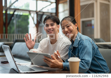 Two cheerful Asian friends, a male and a female, working remotely together from a coffee shop. 118262948