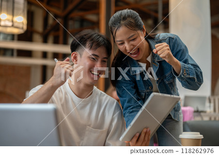 Two cheerful Asian friends are celebrating good news together, looking at a digital tablet. 118262976