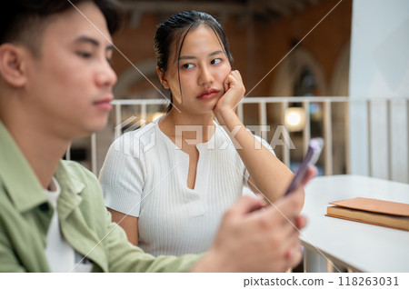 An unhappy Asian woman is feeling jealous, looking at her boyfriend while he is texting on his phone An unhappy Asian woman is feeling jealous, looking at her boyfriend while he is texting on his phone 118263031