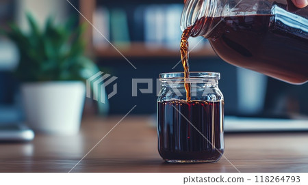 Freshly Brewed Coffee Being Poured into a Glass Jar on a Wooden Table with Green Plant in Background 118264793