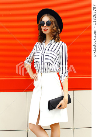 Beautiful young woman model wearing white striped shirt, black handbag clutch, round hat, skirt in the city 118265797