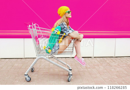 cool young woman sitting in trolley cart with skateboard over colorful pink background 118266285