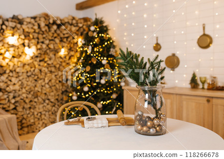 Christmas tree is lit up with lights in front of wall of wood. Vase with green plant on a table Christmas tree is lit up with lights in front of wall of wood. Vase with green plant on a table 118266678