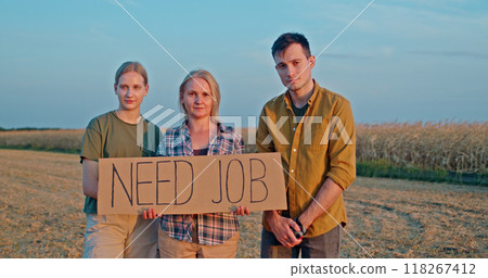 A farming family of three holds a cardboard sign that says Need Job while standing in a cornfield. The group looks concerned, seeking help during harvest season. 118267412