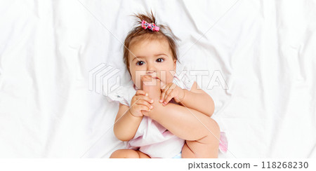 With playful smile, baby girl puts her foot in her mouth, showing her adorable innocence while lying comfortably on bed. With playful smile, baby girl puts her foot in her mouth, showing her adorable innocence while lying comfortably on bed. 118268230