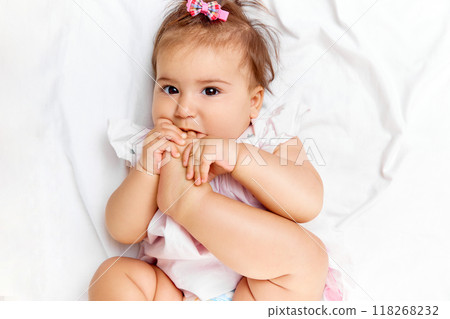 With playful smile, baby girl puts her foot in her mouth, showing her adorable innocence while lying comfortably on bed. With playful smile, baby girl puts her foot in her mouth, showing her adorable innocence while lying comfortably on bed. 118268232