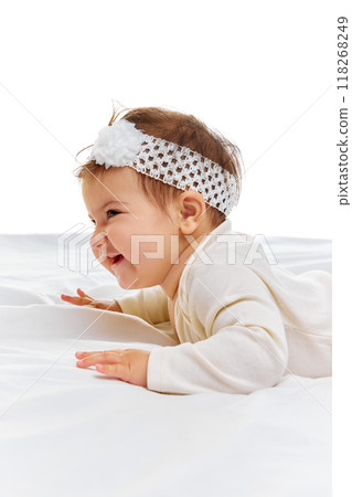 Side view photo of darling little cherub, dressed in simple white onesie and headband, full of curiosity, lying comfortably on white surface. Side view photo of darling little cherub, dressed in simple white onesie and headband, full of curiosity, lying comfortably on white surface. 118268249