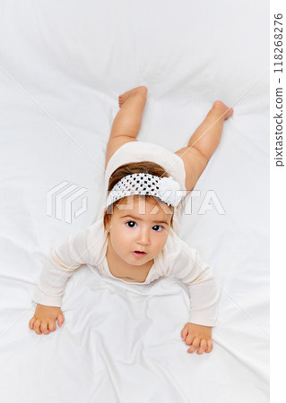 Young child in white onesie lies flat on her stomach, gazing intently at camera against white studio background. Young child in white onesie lies flat on her stomach, gazing intently at camera against white studio background. 118268276
