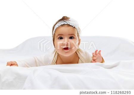 Little baby in white onesie and headband looks curiously at camera, crawling on soft white bed with focused expression. Little baby in white onesie and headband looks curiously at camera, crawling on soft white bed with focused expression. 118268293