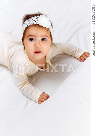 Young child with flower headband and outfit looks directly camera, her big eyes wide, while lying on bright white bed. Young child with flower headband and outfit looks directly camera, her big eyes wide, while lying on bright white bed. 118268299