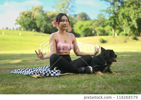Calm young woman sitting in Lotus Pose meditating next to pet dog outdoors 118268579