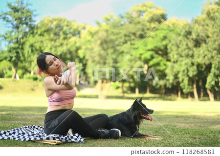 Young woman sitting on a blanket in a park with her border collie dog and stretching her arms after workout 118268581
