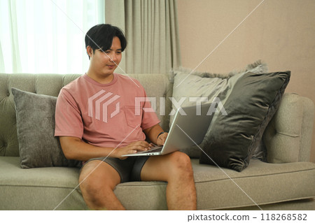 Smiling young asian man in casual clothing using laptop on comfortable sofa at home 118268582